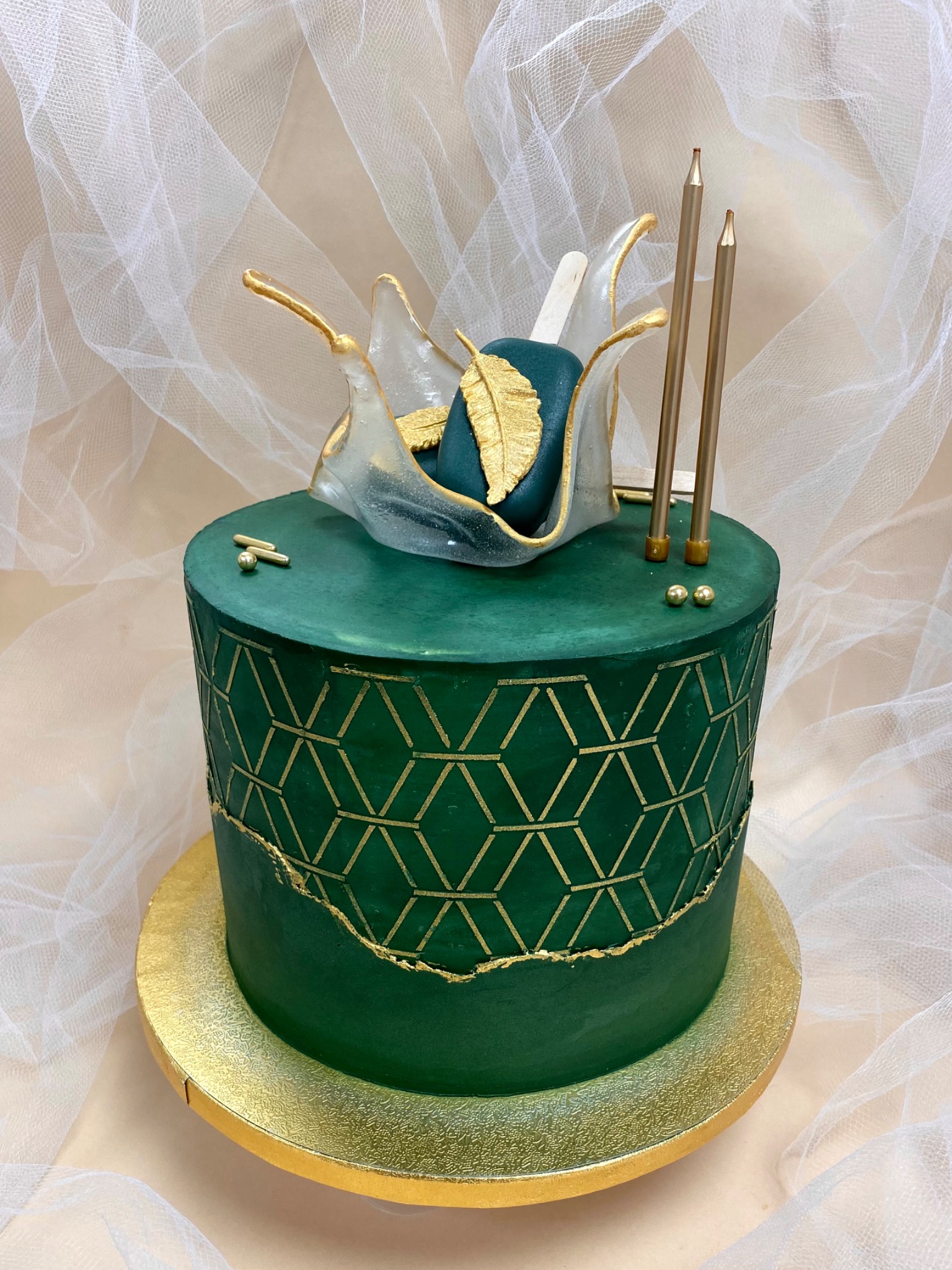 Elegancki tort urodzinowy w odcieniach zieleni z geometrycznym złotym wzorem, ozdobiony jadalną dekoracją przypominającą kryształową fontannę z zielonym elementem i złotym piórkiem oraz trzema...