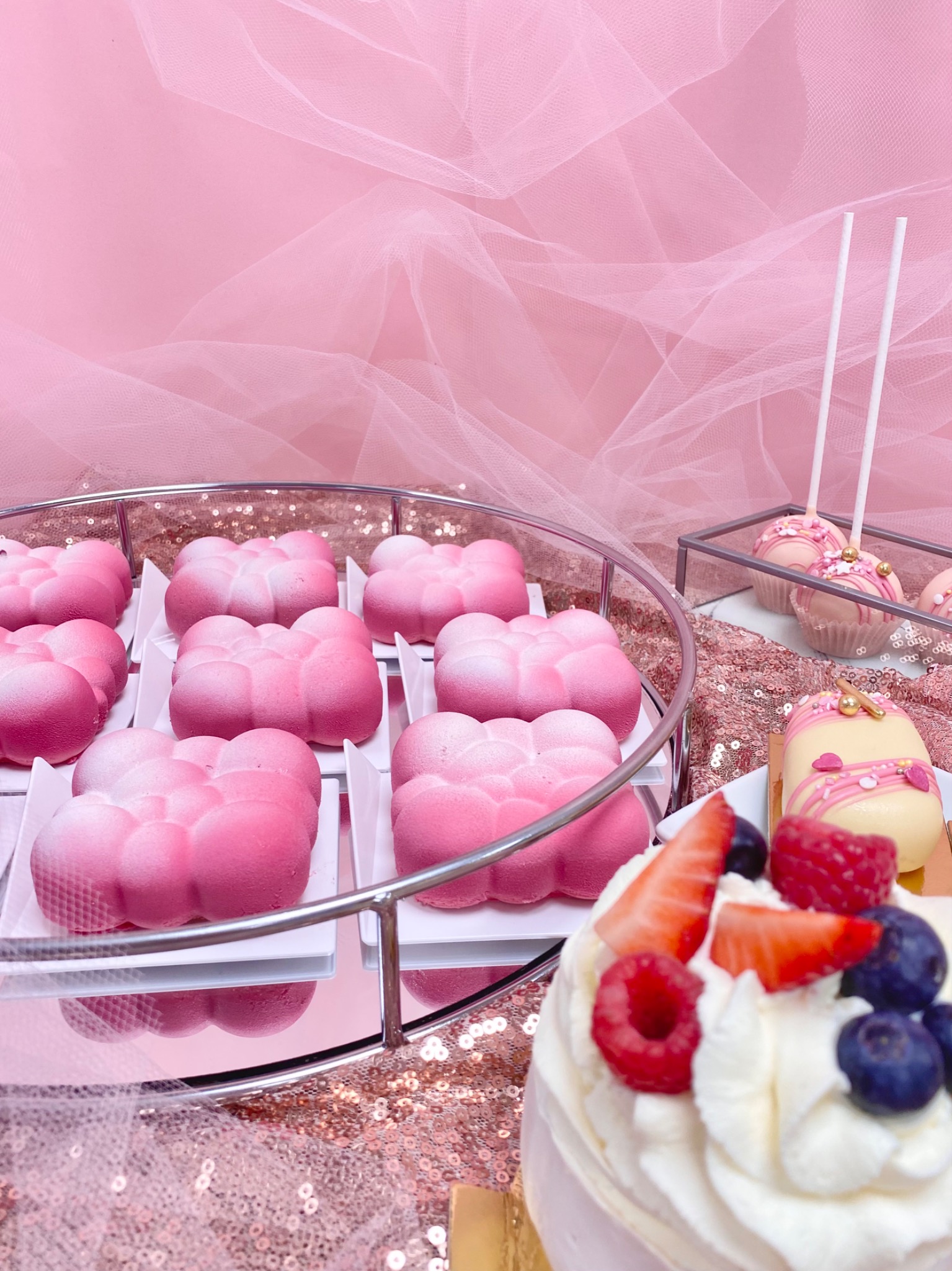 Różowe desery w kształcie chmurek na srebrnej tacy, cake pops i mini torcik z owocami na tle różowego tiulu i cekinów.