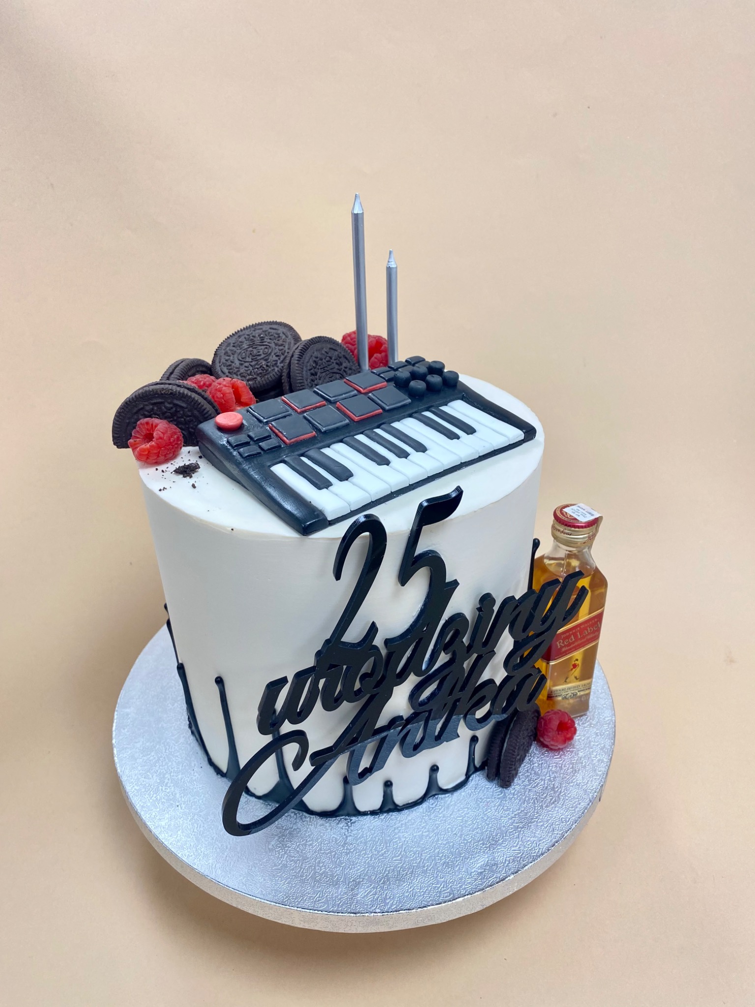 Tort urodzinowy z dekoracjami: miniaturowy syntezator, ciasteczka Oreo, maliny, buteleczka whisky Johnnie Walker Red Label i czarny napis '25 urodziny Antka', całość na srebrnej paterze.