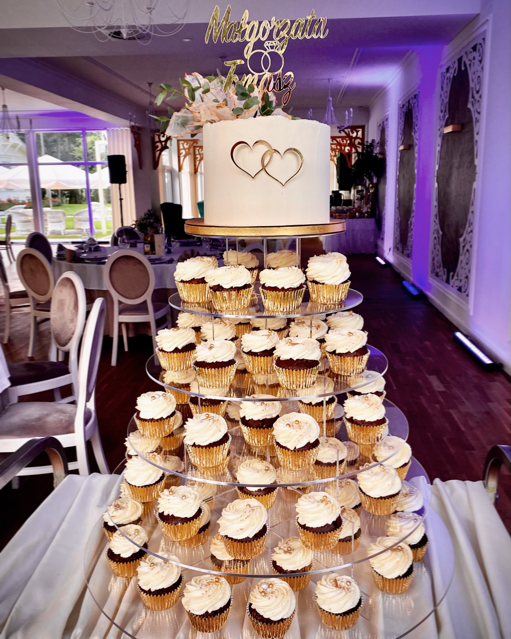 Wielopoziomowa konstrukcja z babeczek z kremem i złotymi papilotkami, zwieńczona tortem z dekoracją imion Małgorzata i Tomasz oraz symbolami obrączek, w tle sala bankietowa z nakrytymi stołami.