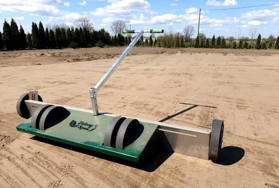 Niwelator do trawy z rolki marki Zielony Spaw na przygotowanym terenie, widoczne drzewa w tle.