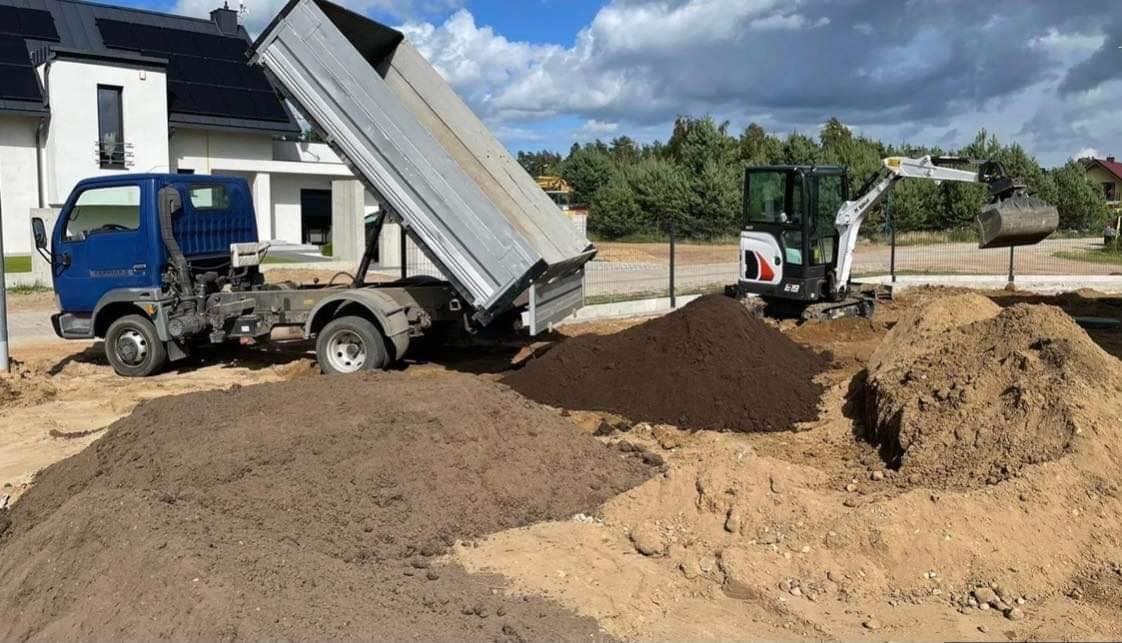 Niebieska ciężarówka z wywrotką i biała minikoparka Bobcat na placu budowy z pryzmami ziemi różnego koloru, w tle dom z panelami słonecznymi i drzewa.