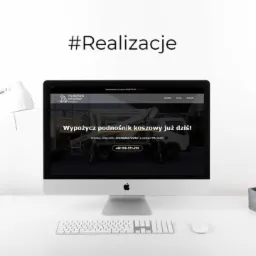 Ekran komputera iMac wyświetlający stronę internetową firmy oferującej wynajem podnośników koszowych, obok lampka biurkowa i kaktus w szarej doniczce, na białym biurku.