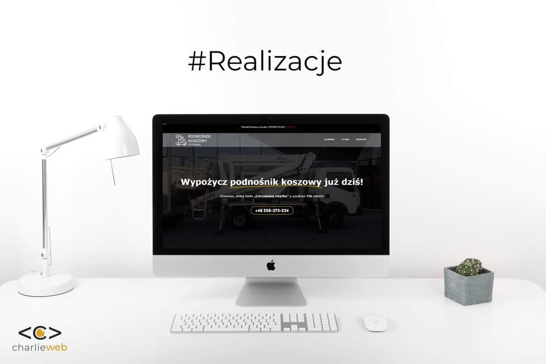 Ekran komputera iMac wyświetlający stronę internetową firmy oferującej wynajem podnośników koszowych, obok lampka biurkowa i kaktus w szarej doniczce, na białym biurku.