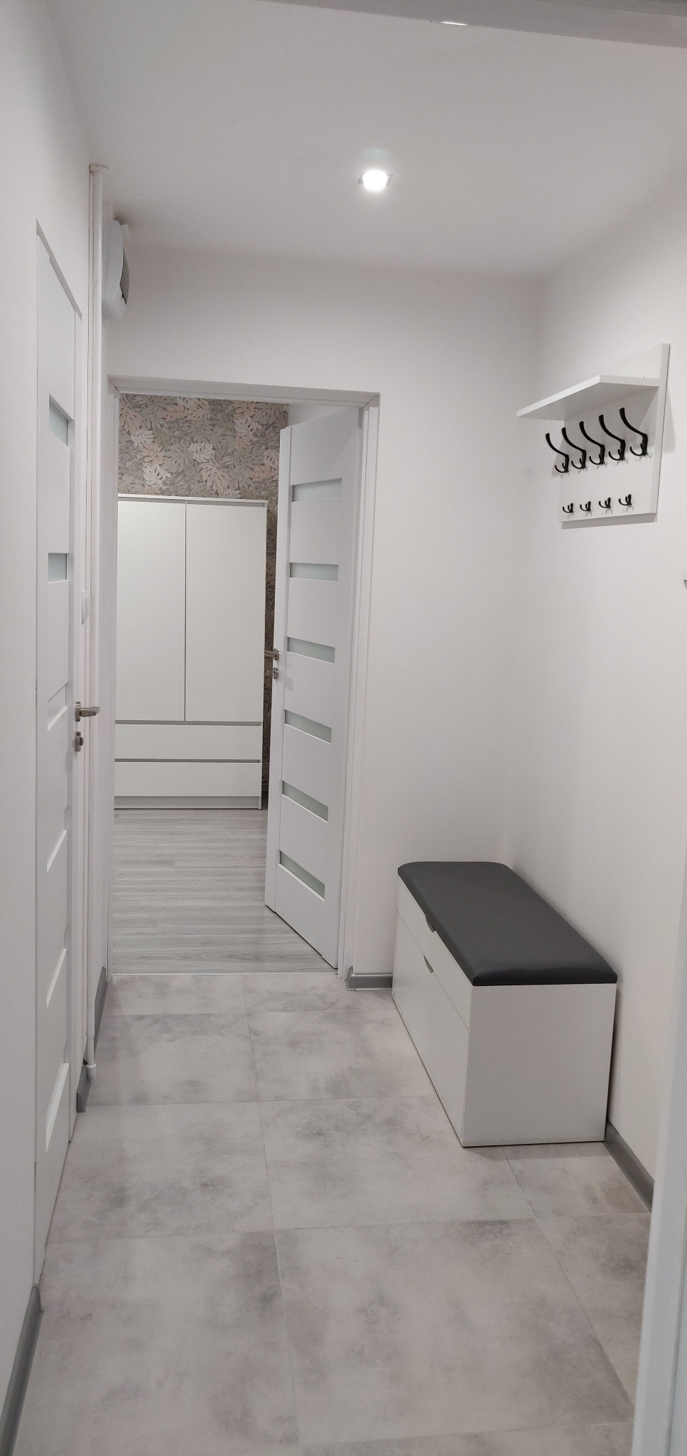 Jasny korytarz z szarymi płytkami podłogowymi, białymi drzwiami z przeszkleniami, szafką z siedziskiem i wieszakiem na ścianie, widok na szafę wnękową z tapetą w liście w tle.