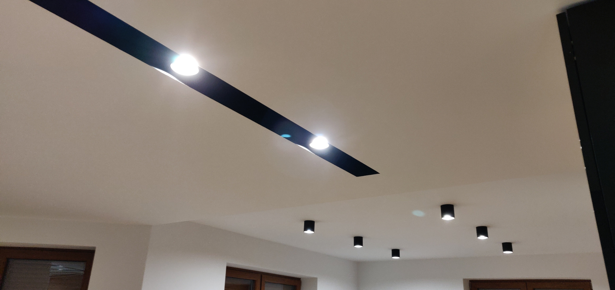 Biały sufit z zamontowanym czarnym profilem oświetleniowym z trzema punktami światła oraz dodatkowymi czarnymi lampami punktowymi. Widoczne fragmenty okien z drewnianą ramą.