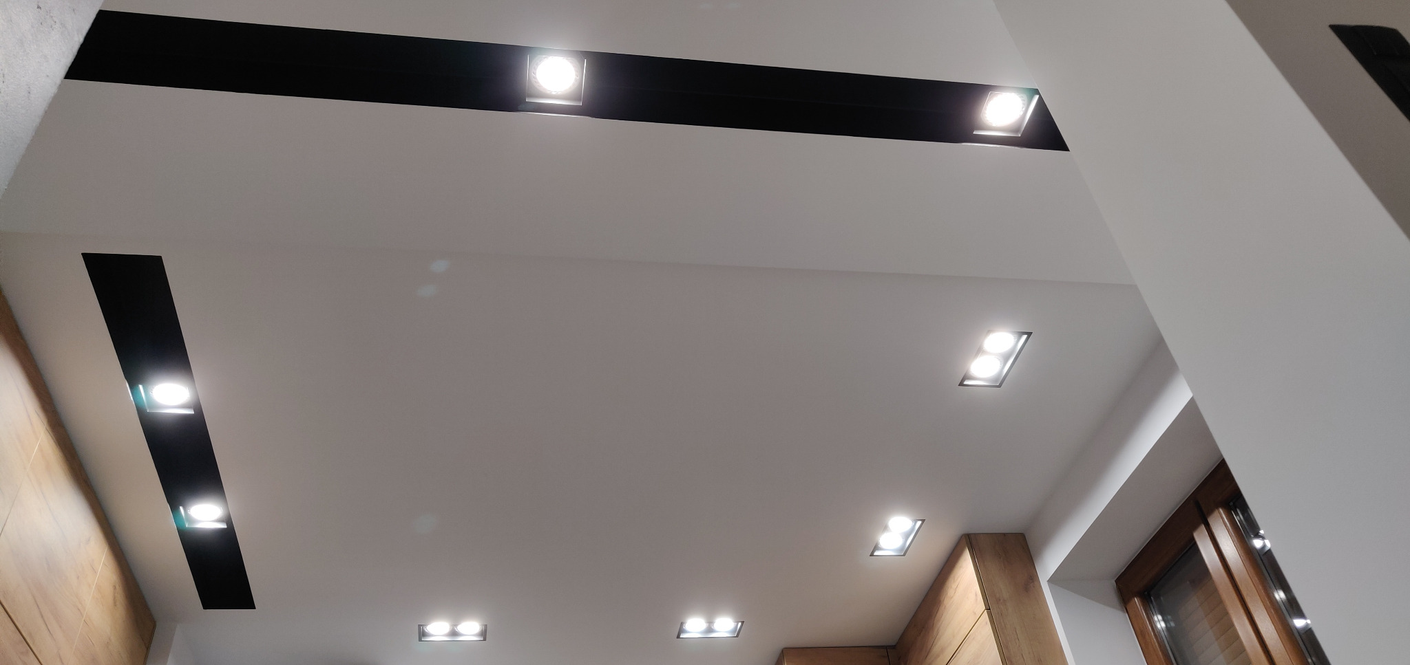 Biały sufit podwieszany z czarnymi pasami i wbudowanymi kwadratowymi lampami LED, oświetlający drewniane wykończenie ściany w narożniku pomieszczenia.