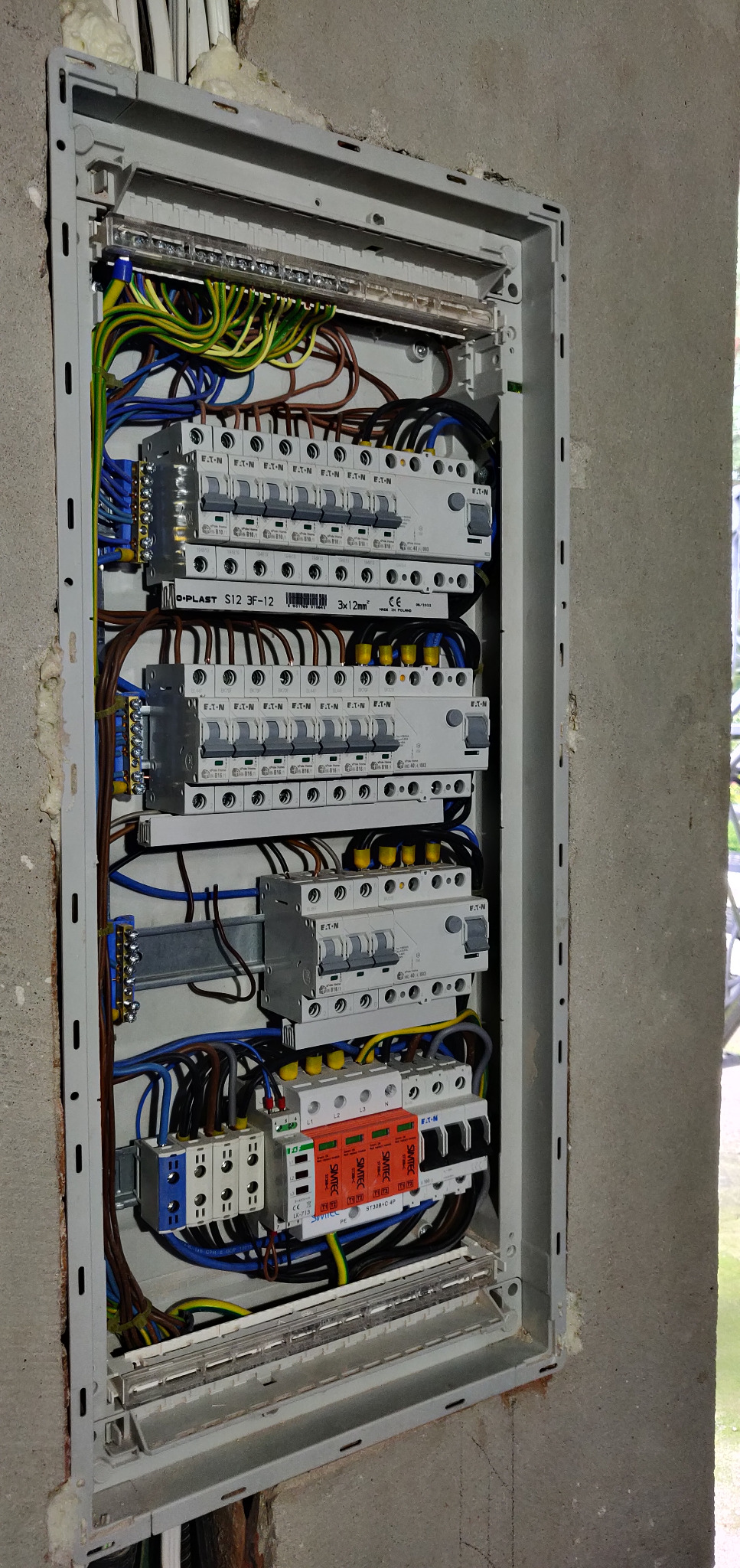 Rozdzielnica elektryczna w szarej obudowie, zamontowana na surowej ścianie, z widocznymi przewodami, bezpiecznikami i zabezpieczeniami przeciwprzepięciowymi marki Simetec, w trakcie montażu lub po...