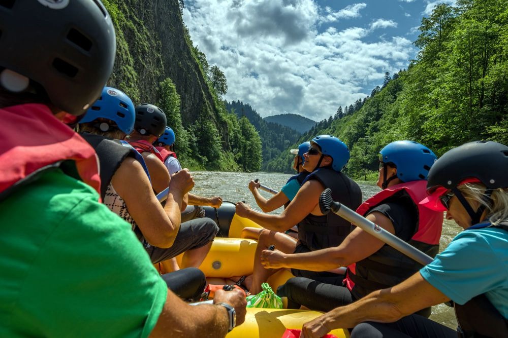 Rafting Dunajcem: grupa w kaskach i kamizelkach płynie pontonem po rzece, w tle skaliste zbocza i zalesione góry Pienińskiego Parku Narodowego, słoneczny dzień.