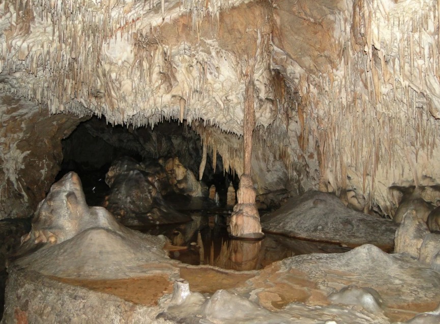 Jaskinia z formacjami naciekowymi: stalaktyty zwisają z sufitu, a w dole woda odbija skały. Oświetlenie podkreśla naturalne piękno podziemnego krajobrazu.