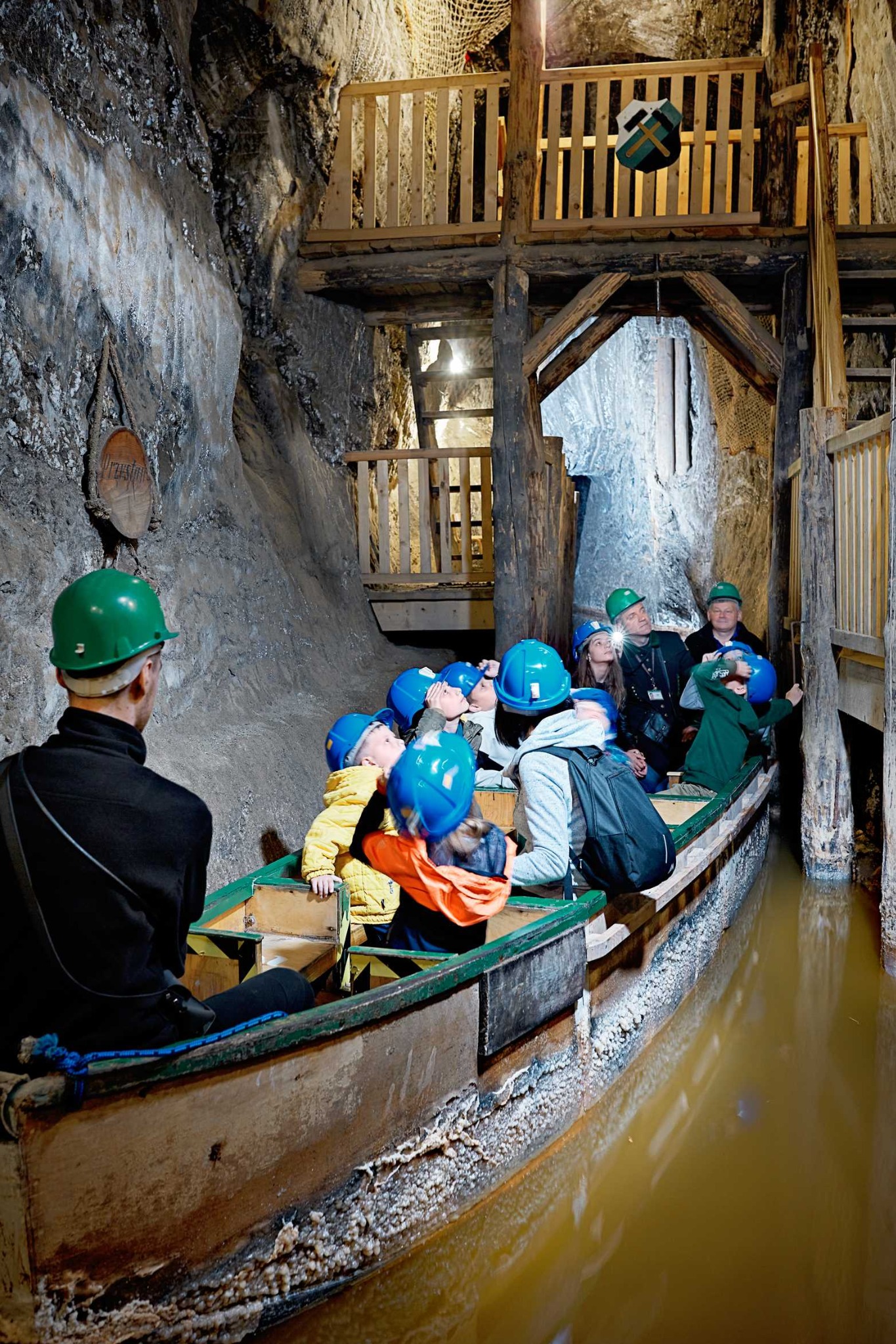 Grupa turystów w kopalni soli płynie łodzią po zalanych korytarzach, w tle drewniana konstrukcja i oświetlone ściany solne. Wszyscy w kaskach.