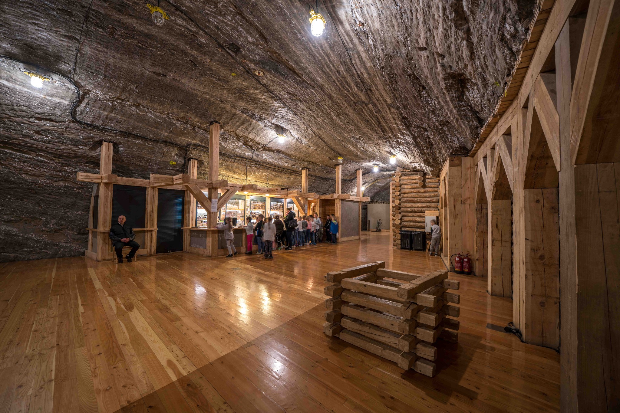 Wnętrze kopalni soli z drewnianą konstrukcją, oświetleniem i grupą zwiedzających w kolejce. Unikalna tekstura ścian i podłogi. Widoczny mężczyzna siedzący na krześle.