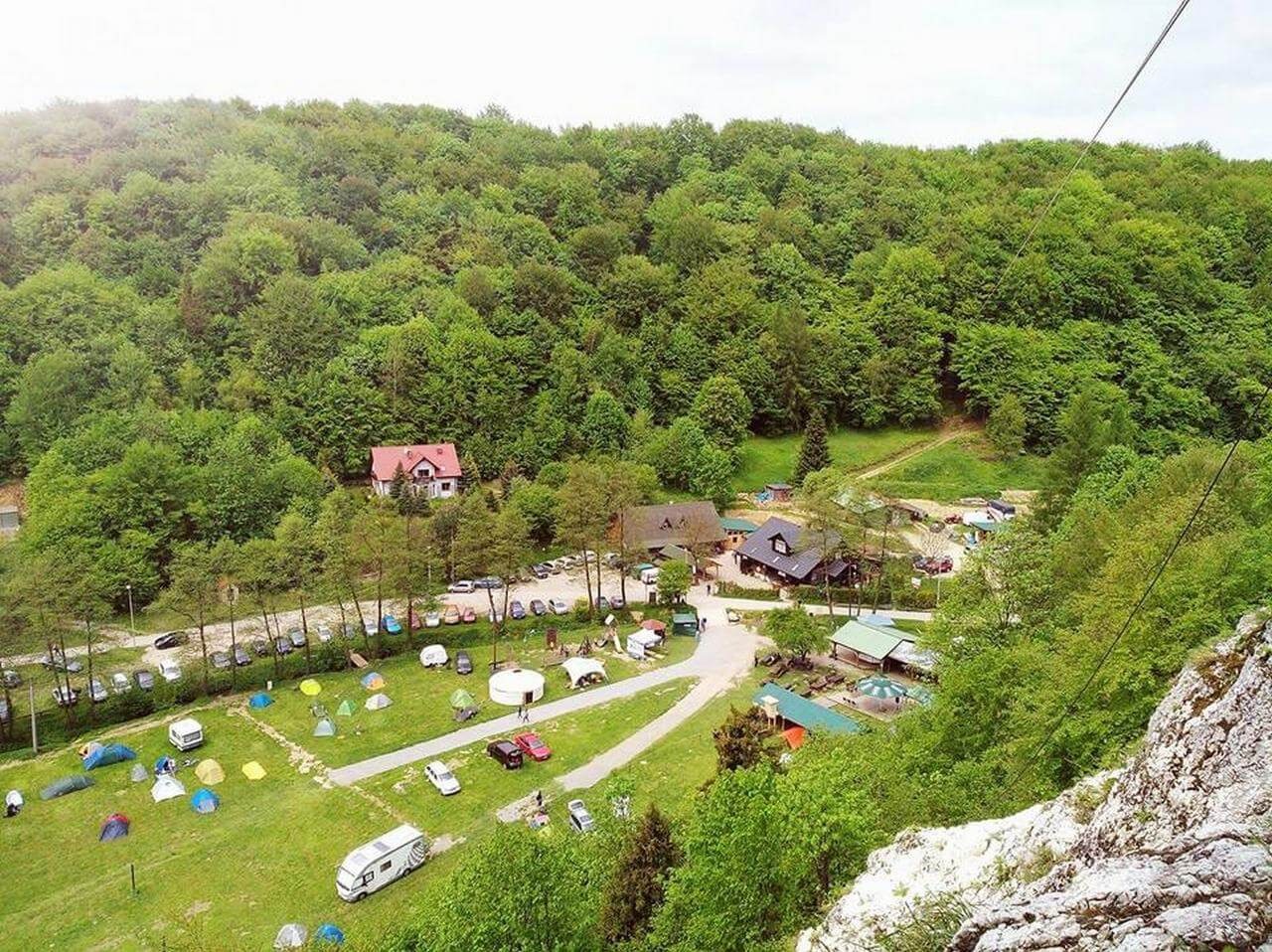 Szeroki kadr z góry na zielony teren rekreacyjny z namiotami, kamperami i budynkami w otoczeniu lasu. Widok z perspektywy skały, w tle gęsty las.