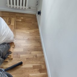 Remont Team - Podłoga z drewnianych paneli w trakcie montażu, widoczny worek z materiałem, odkurzacz i grzejnik.