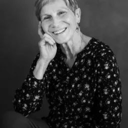 Czarno-biały portret uśmiechniętej starszej kobiety z krótkimi włosami, podpierającej głowę ręką, na ciemnym tle.