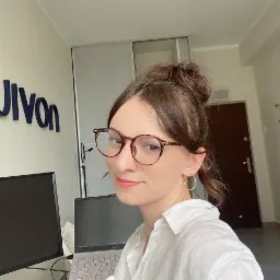 Kobieta w okularach i białej koszuli uśmiecha się w biurze, widoczny laptop i monitor, na ścianie logo firmy.