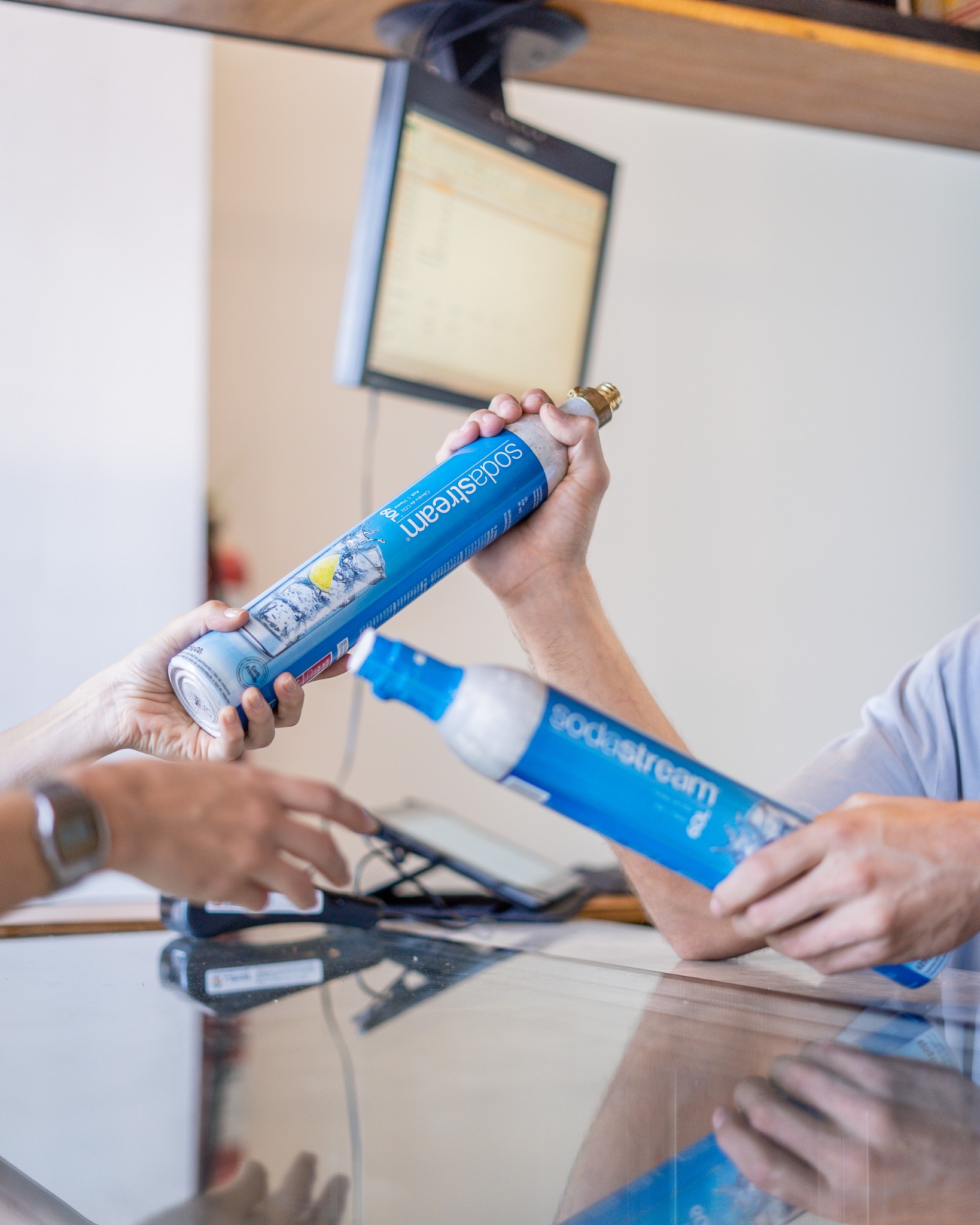 Dwie osoby wymieniają się nabojami CO2 Sodastream przy szklanym blacie, nad którym wisi monitor.