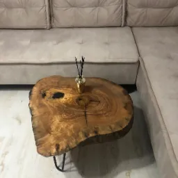 Stolik kawowy z blatem z plastra drewna o nieregularnym kształcie, wsparty na czarnych metalowych nóżkach, obok beżowej, modułowej sofy, z dyfuzorem zapachowym na blacie.