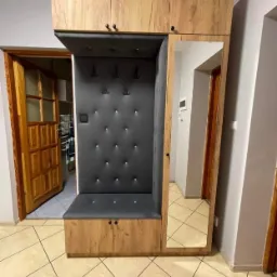 Drewniana zabudowa przedpokoju z tapicerowanym siedziskiem, wieszakami i lustrem, widok na otwarte drzwi do kuchni.