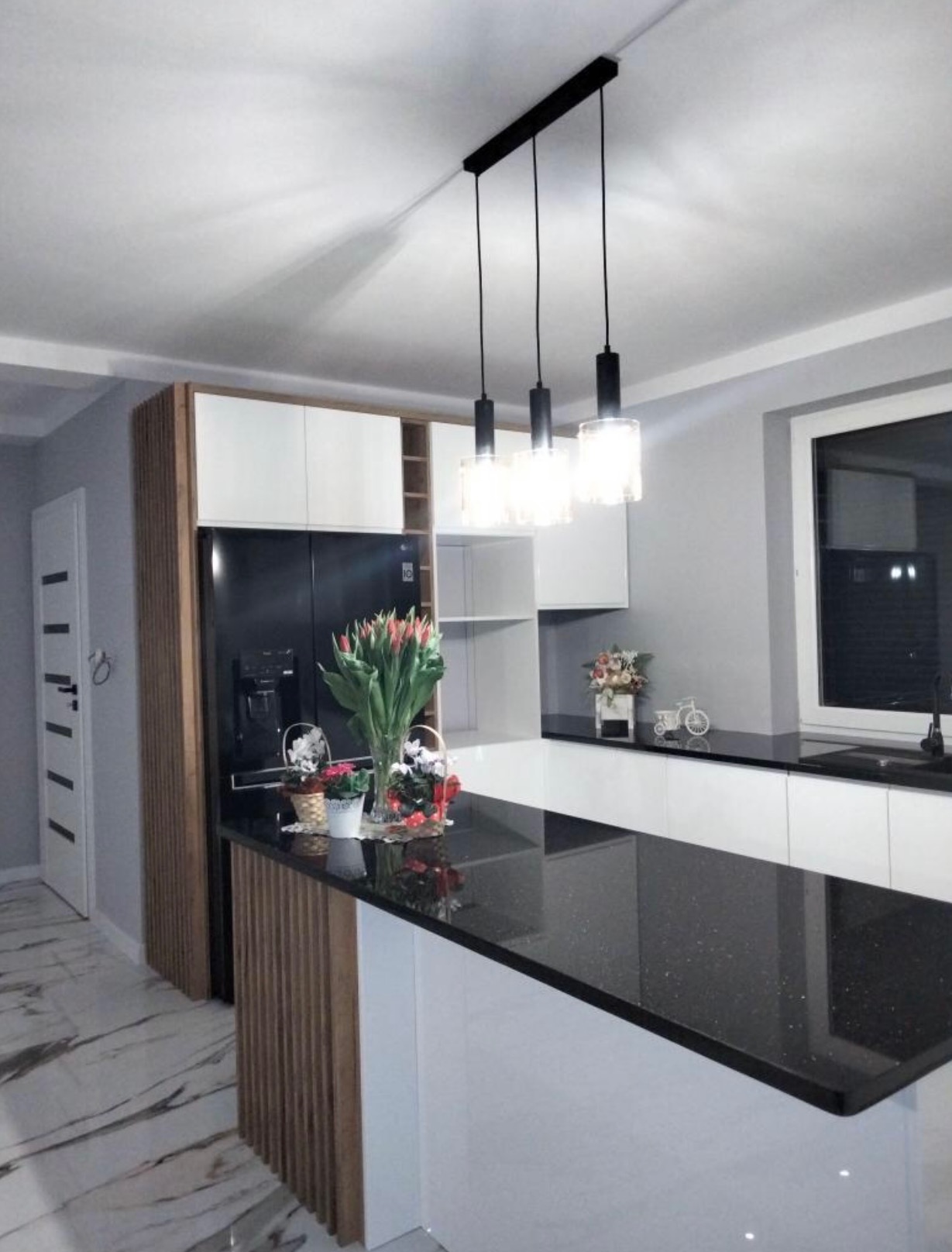 Nowoczesna kuchnia z wyspą, czarny blat z połyskiem, białe szafki, drewniane akcenty i wiszące lampy nad blatem.