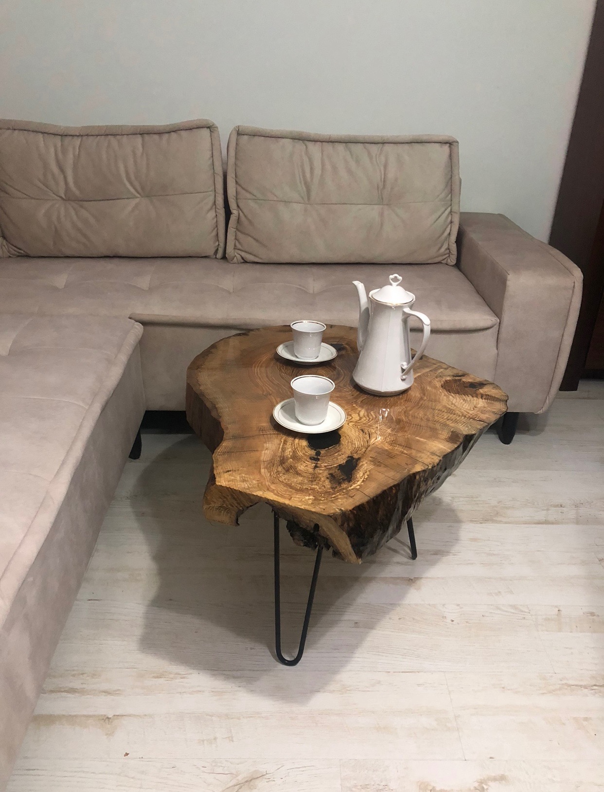 Stolik kawowy z blatem z plastra drewna o nieregularnym kształcie, na metalowych nogach typu hairpin, ustawiony obok beżowej sofy. Na blacie biały dzbanek i dwie filiżanki.