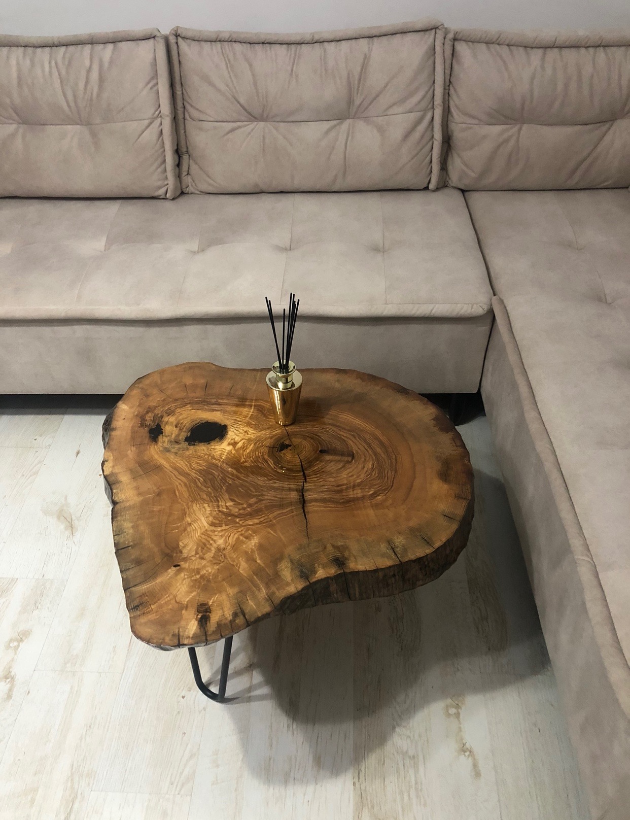 Stolik kawowy z blatem z plastra drewna o nieregularnym kształcie, wsparty na czarnych metalowych nóżkach, obok beżowej, modułowej sofy, z dyfuzorem zapachowym na blacie.