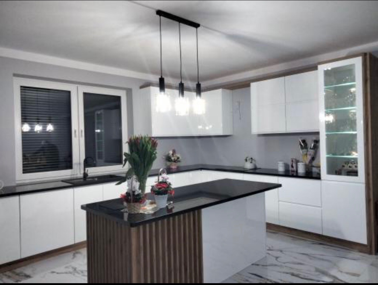 Nowoczesna kuchnia z białymi szafkami, czarnym blatem i wyspą z drewnianymi akcentami, oświetlona wiszącymi lampami i z marmurową podłogą.