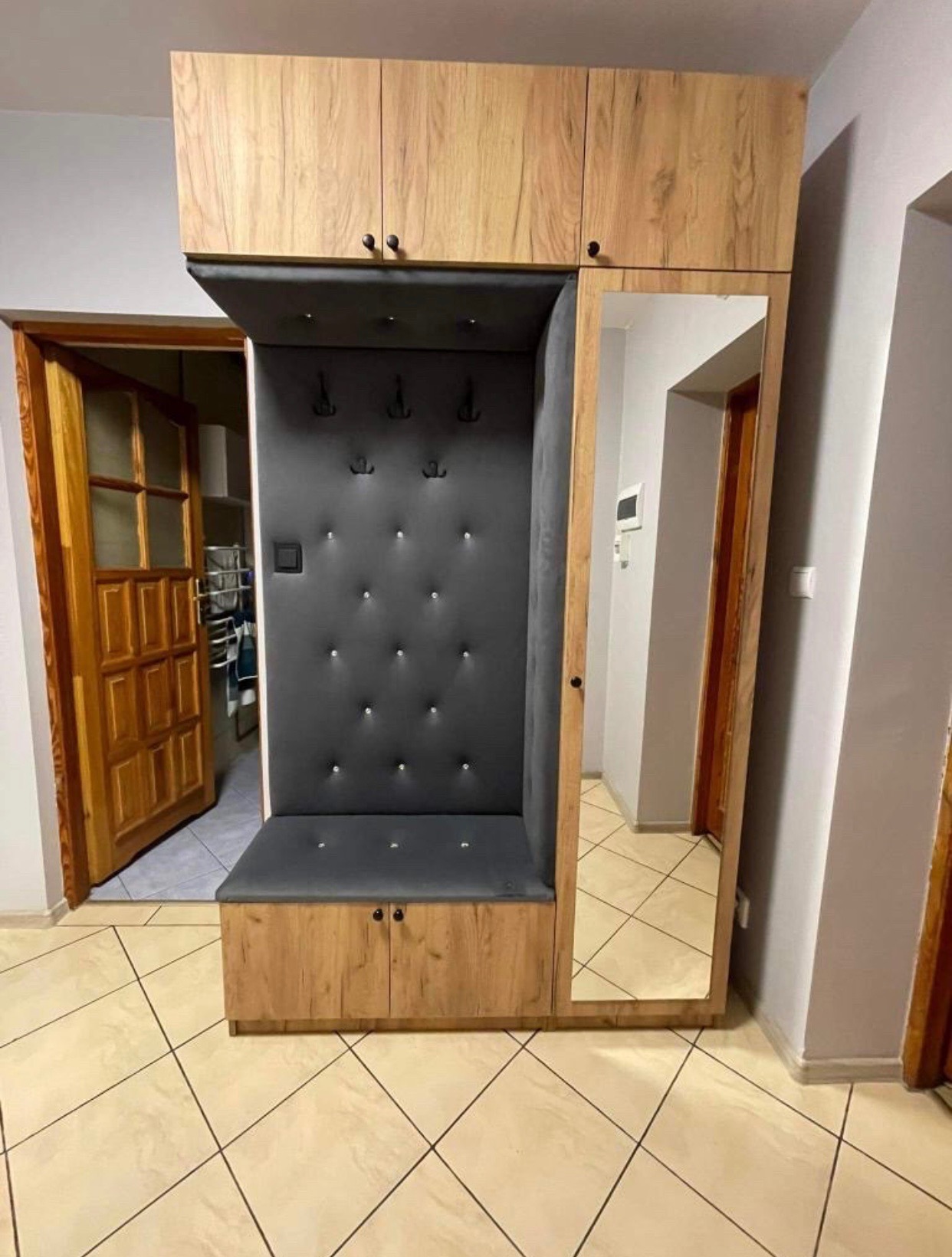 Drewniana zabudowa przedpokoju z tapicerowanym siedziskiem, wieszakami i lustrem, widok na otwarte drzwi do kuchni.