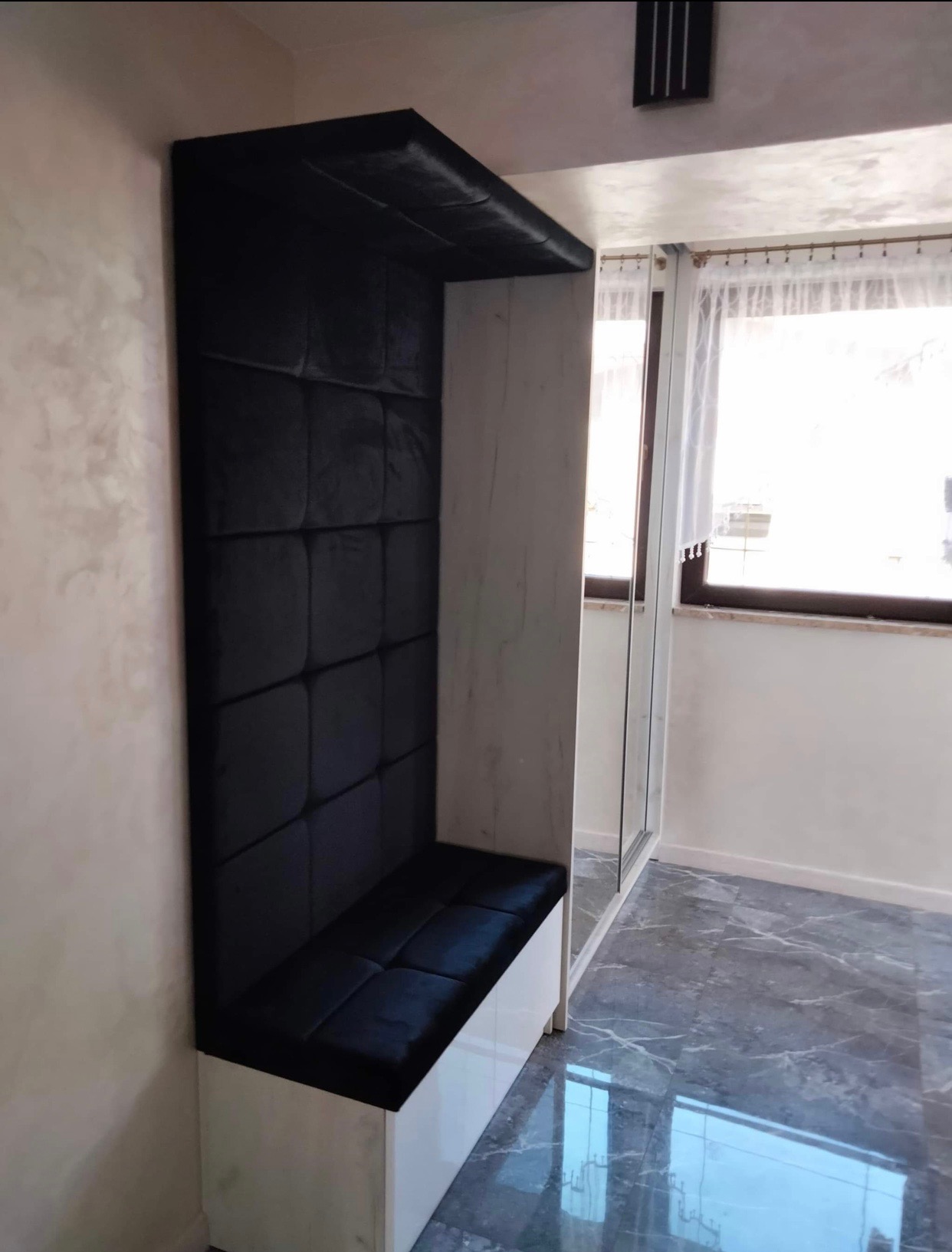 Wnęka z siedziskiem obita czarnym, pikowanym welurem, wkomponowana w białą szafę z lustrzanymi drzwiami, na tle marmurowej podłogi.