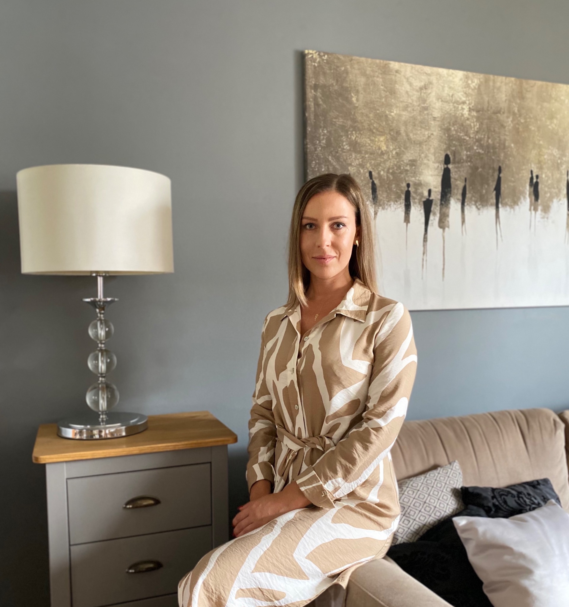 Kobieta siedząca na beżowej sofie w beżowo-białej sukience w zebrę, za nią szara ściana z abstrakcyjnym obrazem w odcieniach złota i czerni oraz stolik nocny z lampką z kryształowymi kulami.