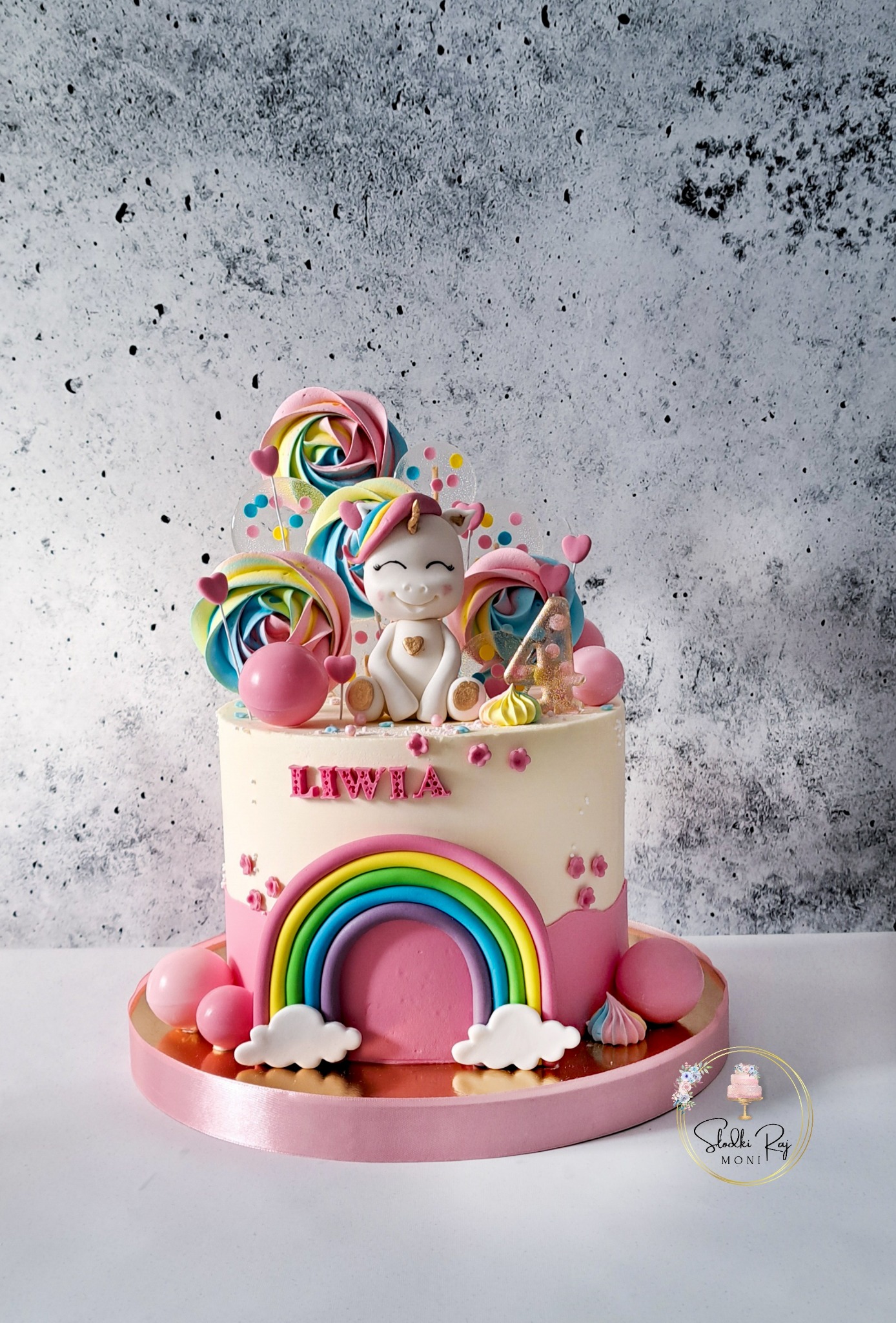 Tort urodzinowy dla dziewczynki Liwii z tęczą, jednorożcem i lizakami w pastelowych kolorach, na tle szarej ściany.