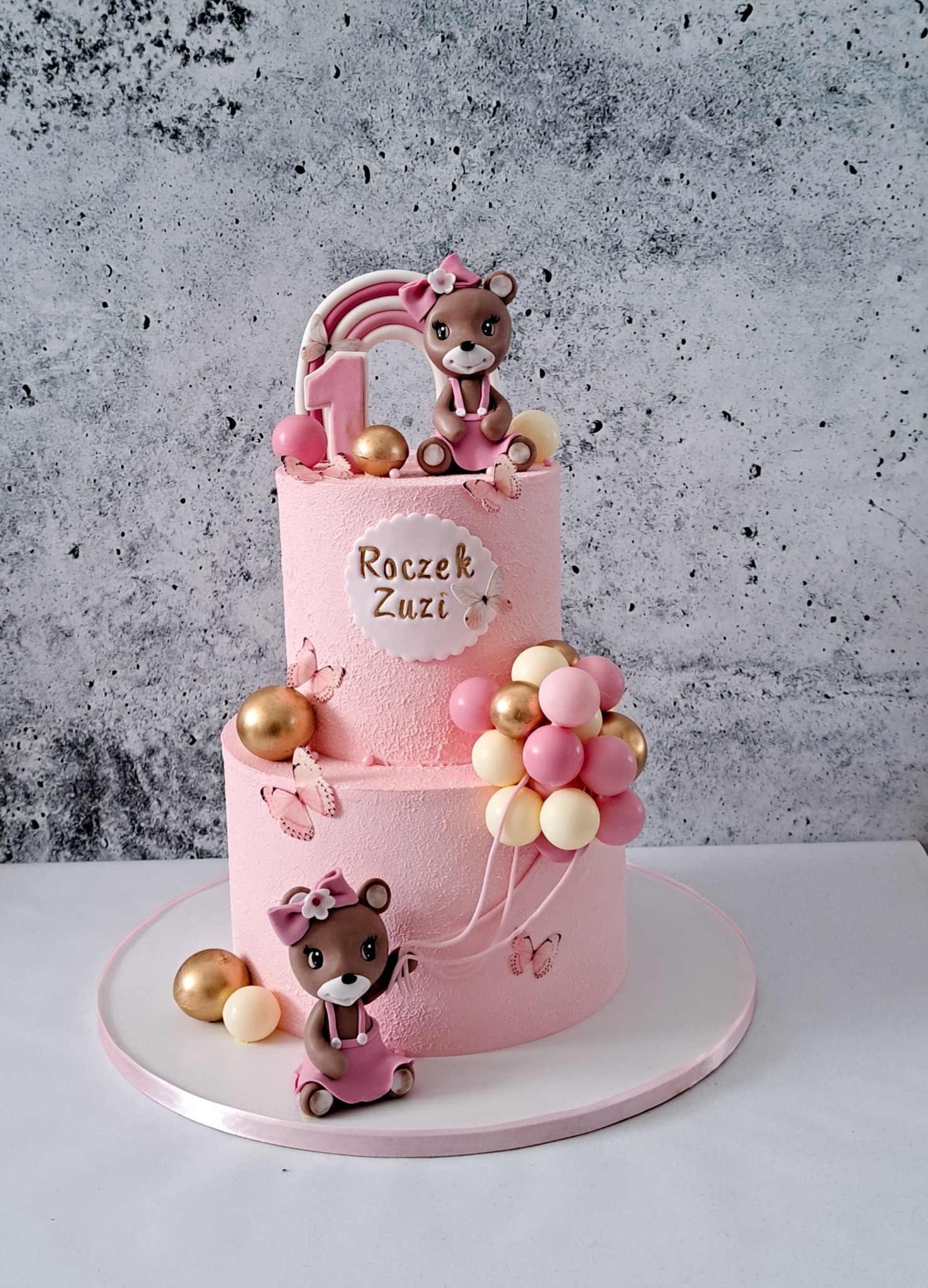 Dwupoziomowy tort w odcieniach różu, zdobiony figurkami misiów z kokardkami, tęczą, cyfrą '1', napisem 'Roczek Zuzi' oraz balonami i motylami.