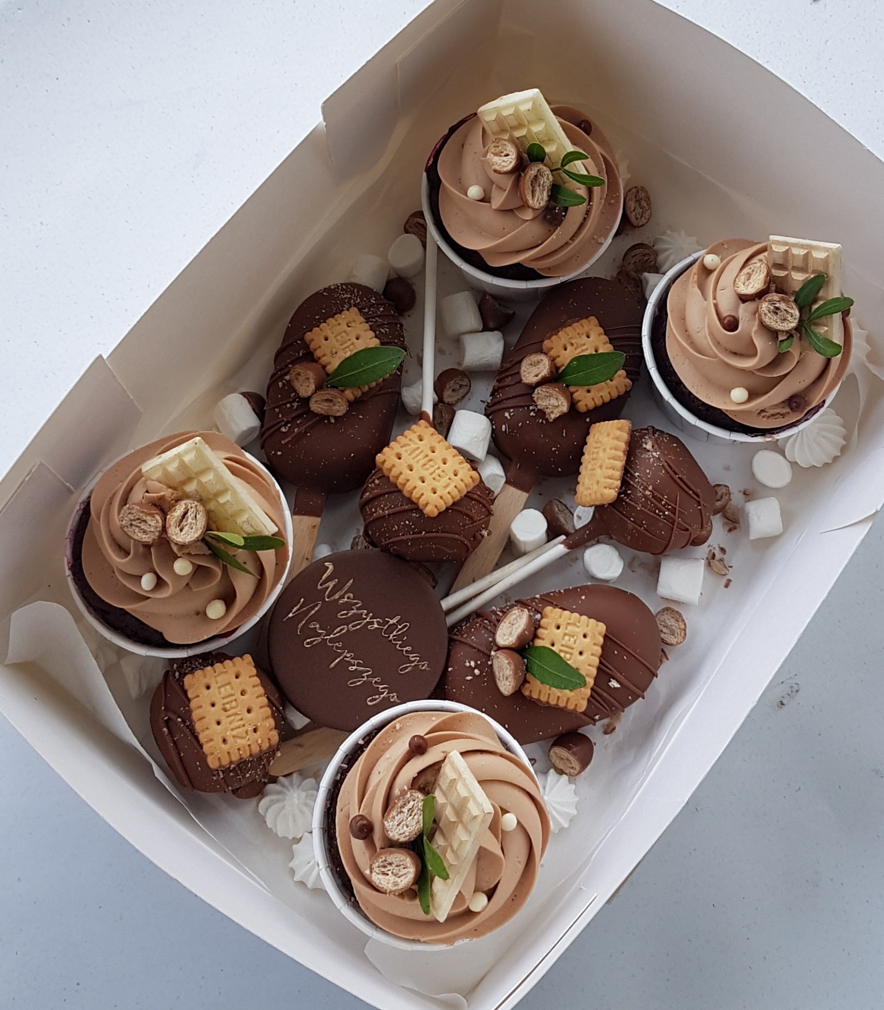 Otwarty karton z czekoladowymi babeczkami z kremem, ciastkami Leibniz i cake pops, udekorowanymi mini bezami i piankami marshmallow.