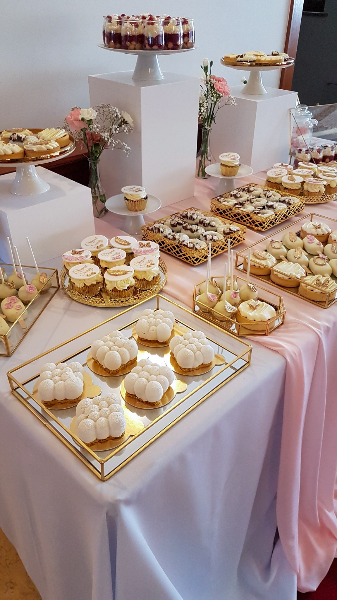 Stół cukierniczy z różnorodnymi deserami: musowe ciastka w kształcie chmurki na lustrzanej tacy, babeczki z personalizowanymi dekoracjami, cake pops z różowymi akcentami, mini tartaletki i desery...