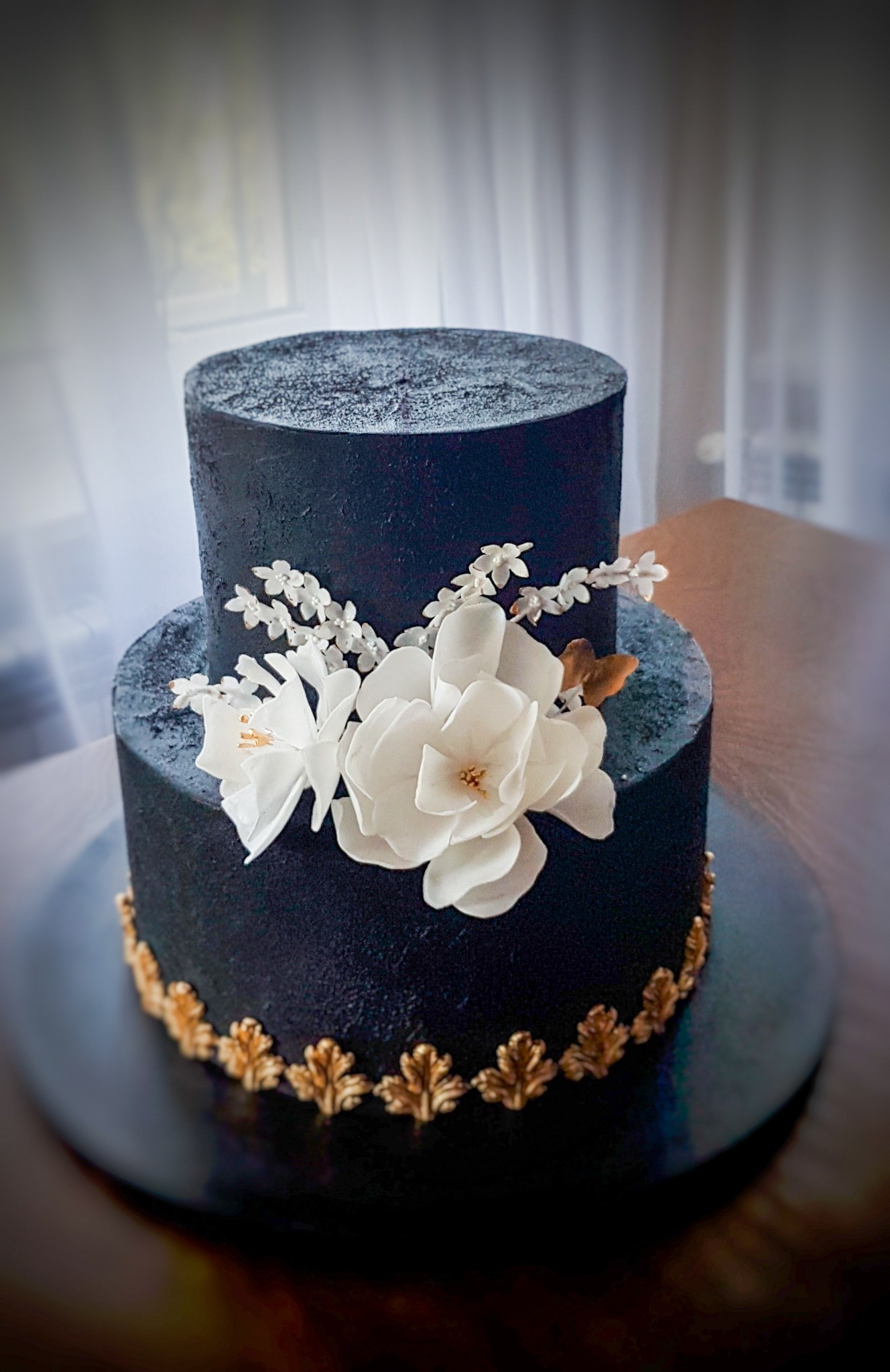 Dwupoziomowy tort w ciemnej tonacji, zdobiony białymi kwiatami i złotymi detalami, umieszczony na ciemnym talerzu.