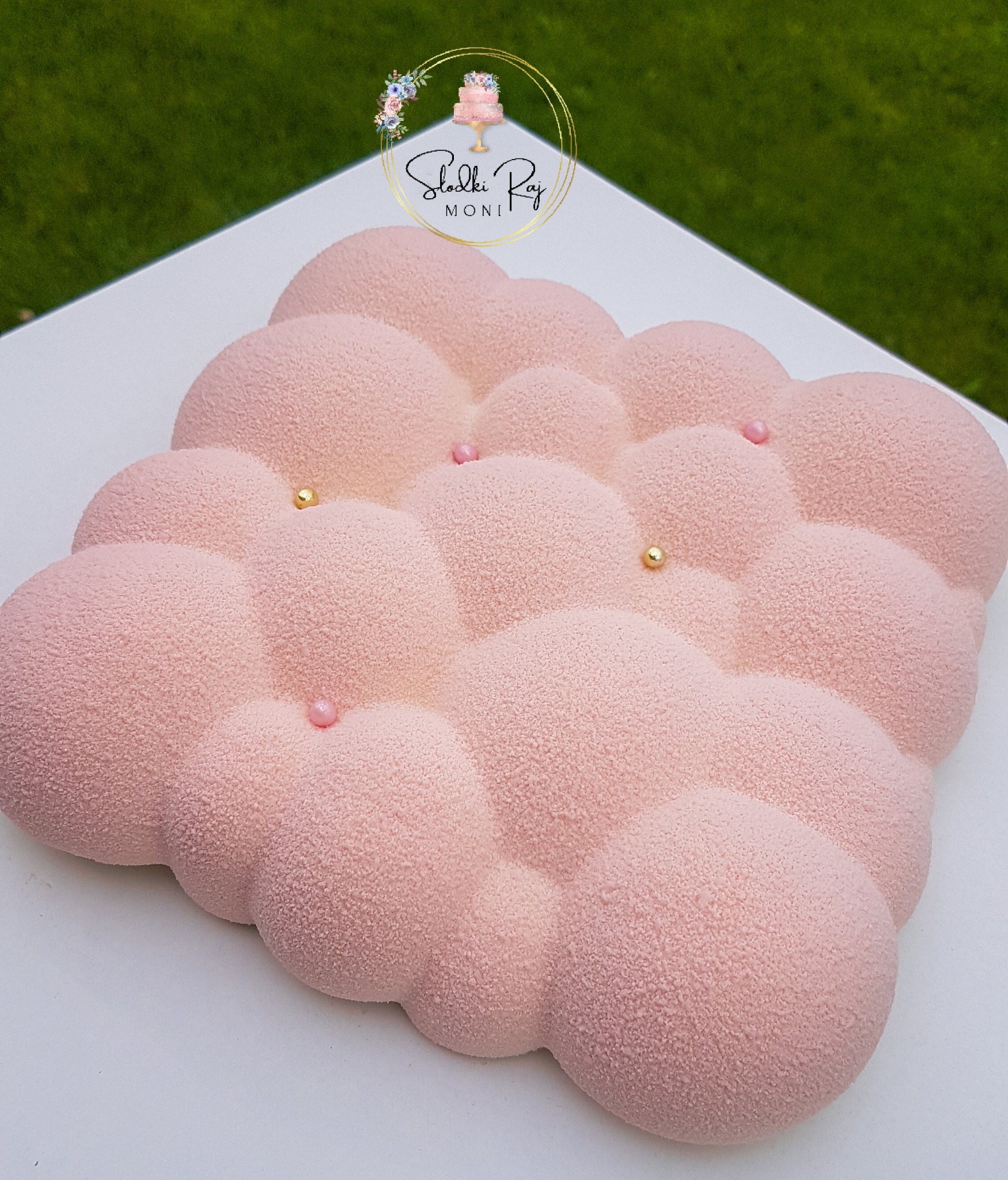 Różowy tort w kształcie chmury z aksamitnym wykończeniem, ozdobiony złotymi i różowymi koralikami na białym tle, z logo firmy w tle.