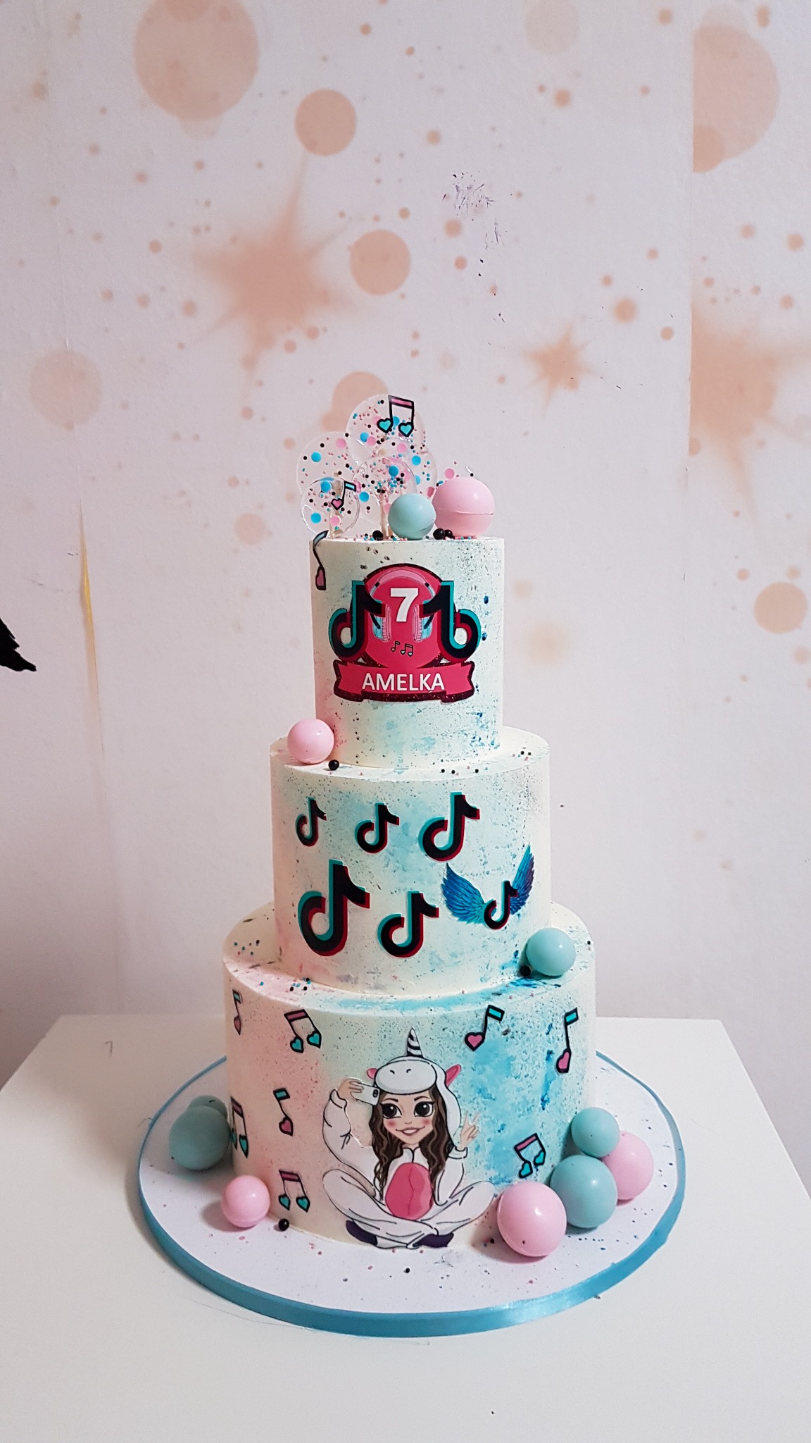 Trzypiętrowy tort urodzinowy w pastelowych kolorach, dekorowany motywami TikTok i postacią w stroju jednorożca, z napisem 'Amelka' i liczbą 7, otoczony dekoracyjnymi kulkami.