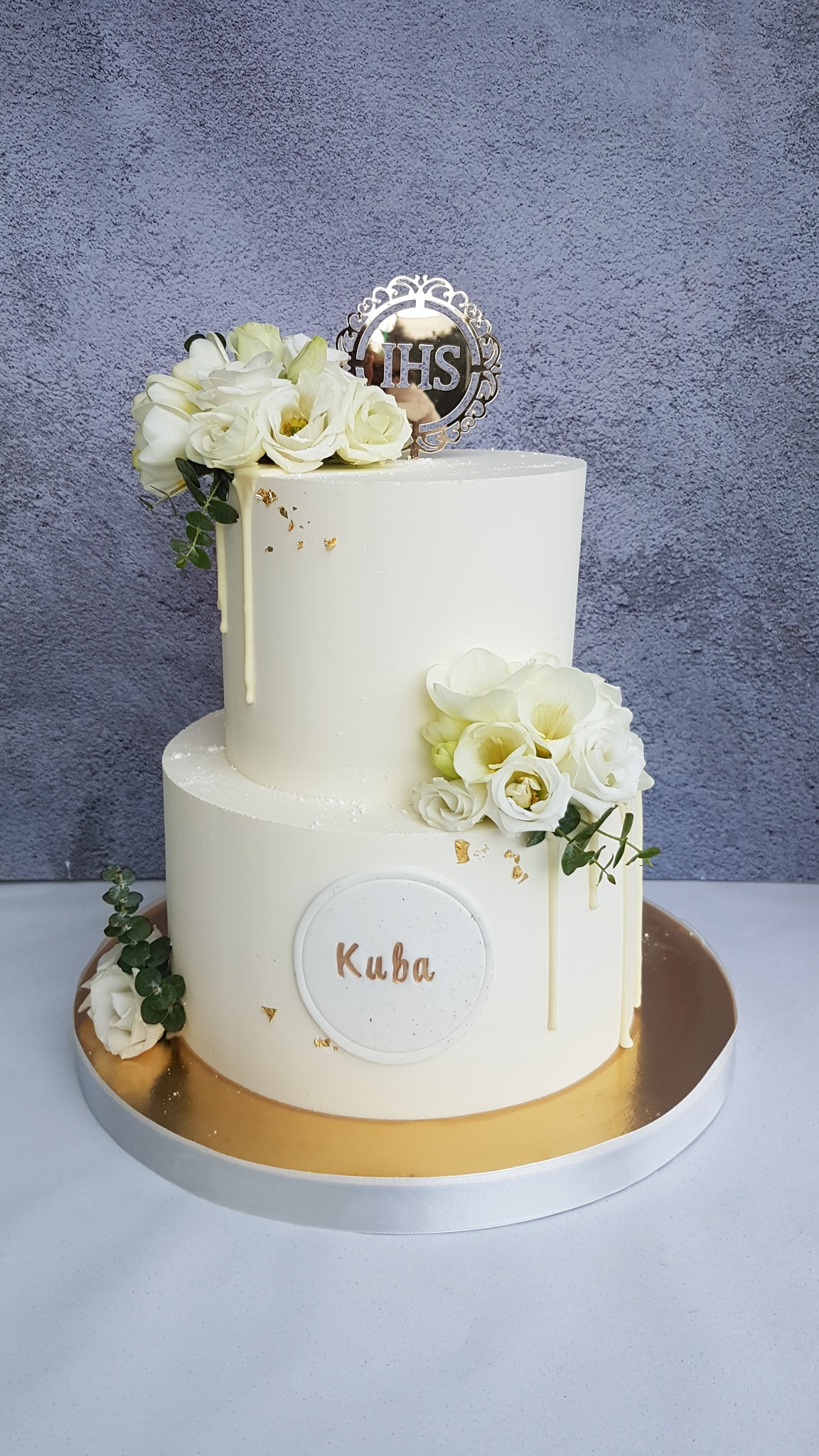Dwupoziomowy tort w białej polewie, zdobiony białymi kwiatami, złotymi płatkami i topperem IHS; na dolnym poziomie okrągła plakietka z napisem 'Kuba'.