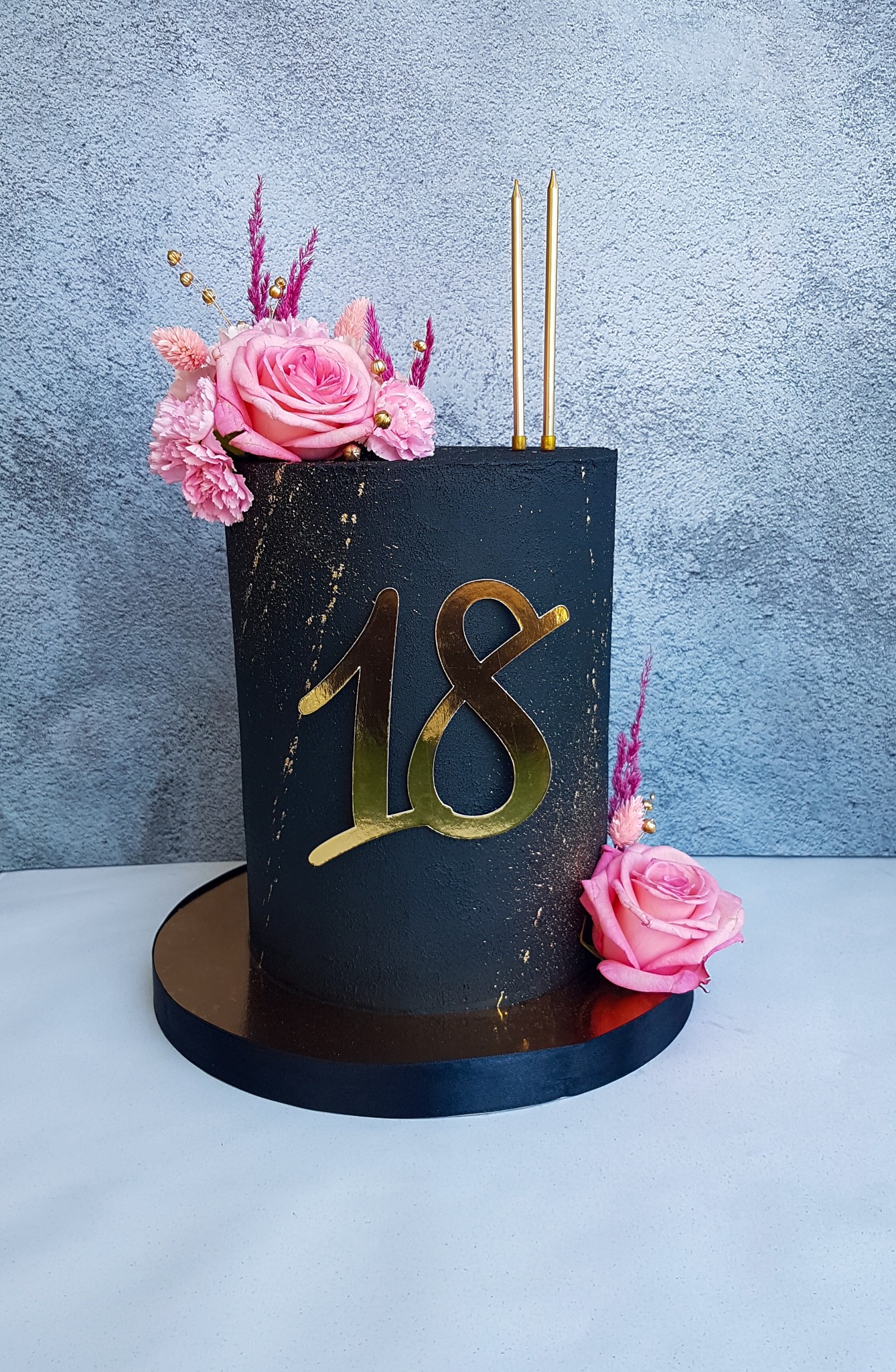 Czarny tort urodzinowy z dekoracją z różowych kwiatów, złotymi akcentami i numerem 18 w kolorze złota, całość na złotym podeście.