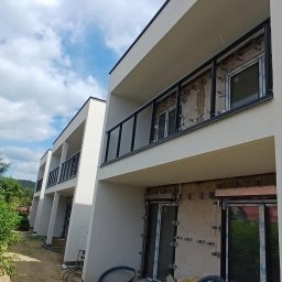 Esterglass - Nowoczesne budynki z jasną elewacją, czarnymi balustradami szklanymi na balkonach piętra. Na parterze widoczne okna w trakcie montażu z izolacją i mocowaniami.