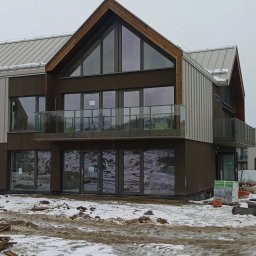 Esterglass - Nowoczesny budynek z ciemną elewacją i drewnianymi szczytami, wyposażony w duże przeszklenia i szklane barierki balkonowe, na tle zaśnieżonego placu budowy.