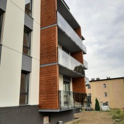 Esterglass - Nowoczesny budynek mieszkalny z elewacją z jasnego tynku, ciemnoszarego cokołu i drewnianych paneli. Balkony posiadają przezroczyste szklane balustrady.