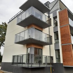 Esterglass - Nowoczesny budynek mieszkalny z wielopoziomowymi balkonami. Balustrady wykonane ze szkła i ciemnego metalu, elewacja w odcieniach beżu i szarości z drewnianymi panelami.