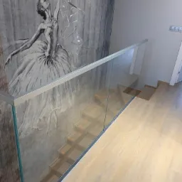 Szklana balustrada schodowa z aluminiowymi profilami, widok z góry na jasne drewniane schody i dekoracyjną ścianę z motywem baletnicy.