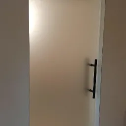 Matowe drzwi z czarną, prostą klamką, zamontowane w jasnej ścianie, widok z bliska na detale wykończenia.