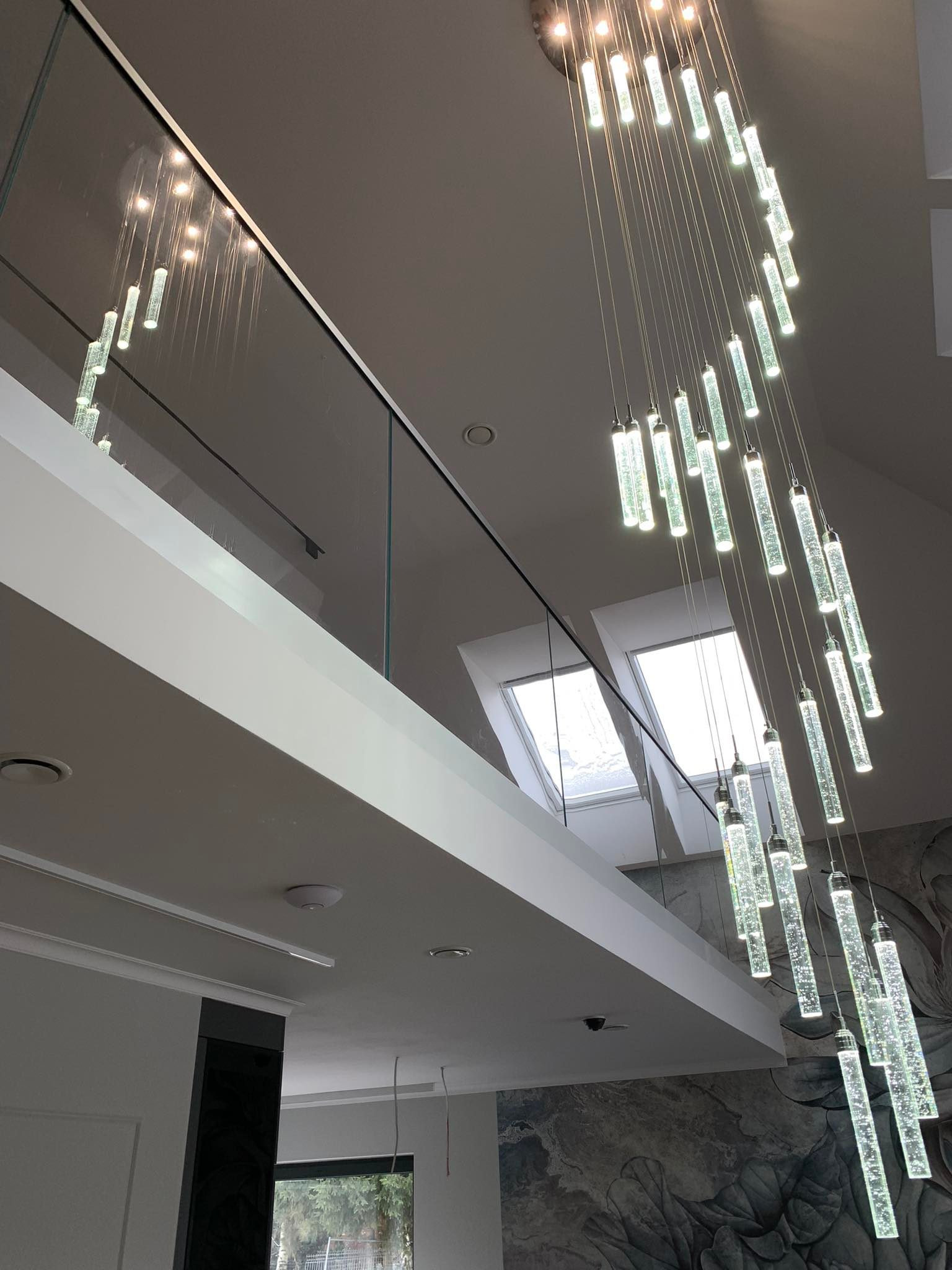 Szklana balustrada na antresoli z widokiem na okna dachowe i dekoracyjny żyrandol z podświetlanych szklanych rurek.