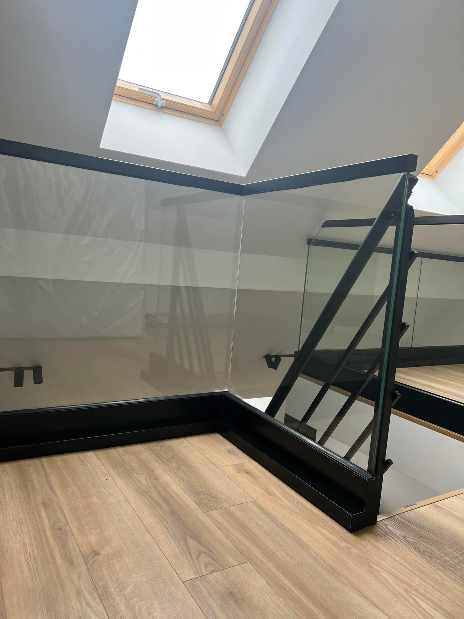 Szklana balustrada z czarnymi metalowymi elementami mocującymi, widoczna z bliska na tle drewnianej podłogi i okna dachowego.