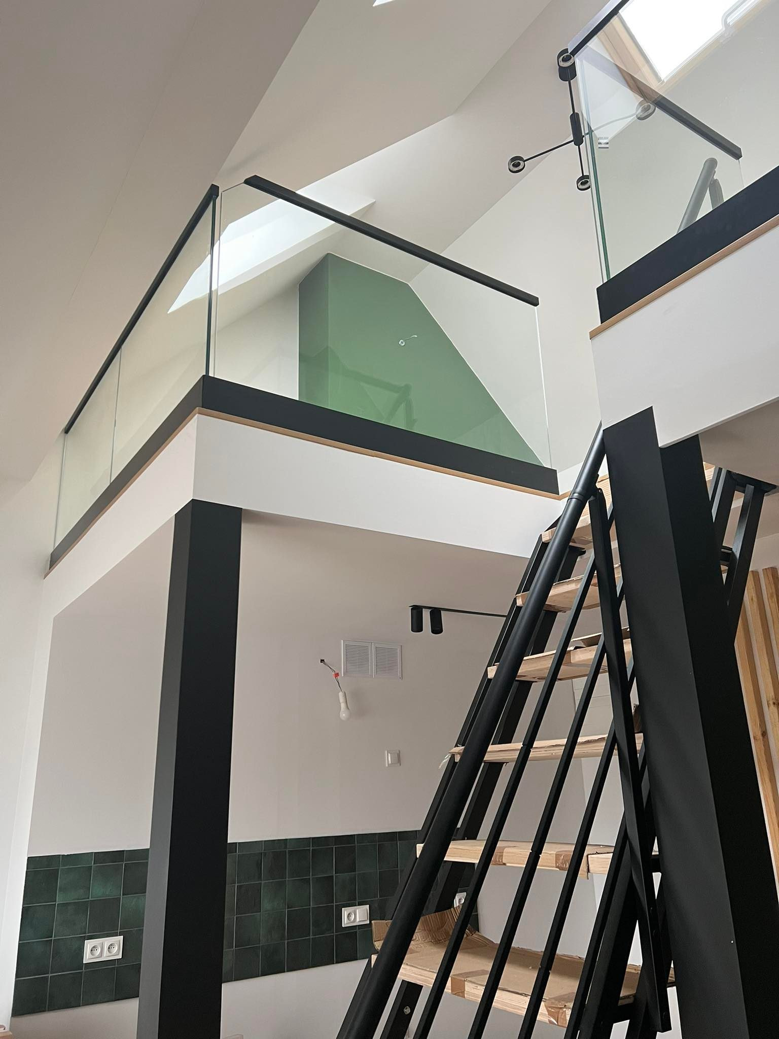 Wnętrze domu z antresolą zabezpieczoną szkłem z czarną ramą, widoczne schody z drewnianymi stopniami i czarną metalową konstrukcją, tło stanowią białe ściany z fragmentem zielonej ściany.