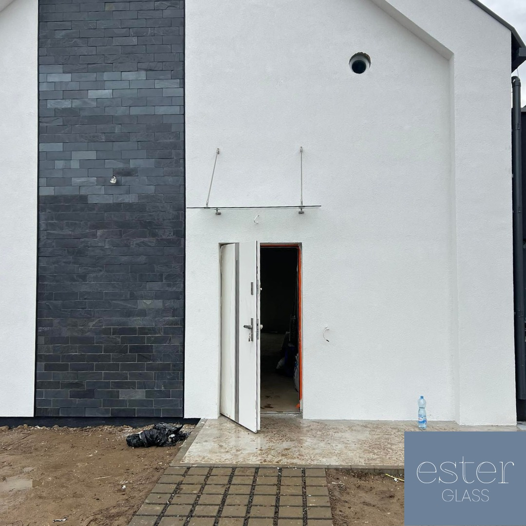 Wejście do budynku z przeszklonym zadaszeniem nad drzwiami, biała elewacja i element dekoracyjny z ciemnego kamienia, częściowo otwarte białe drzwi.