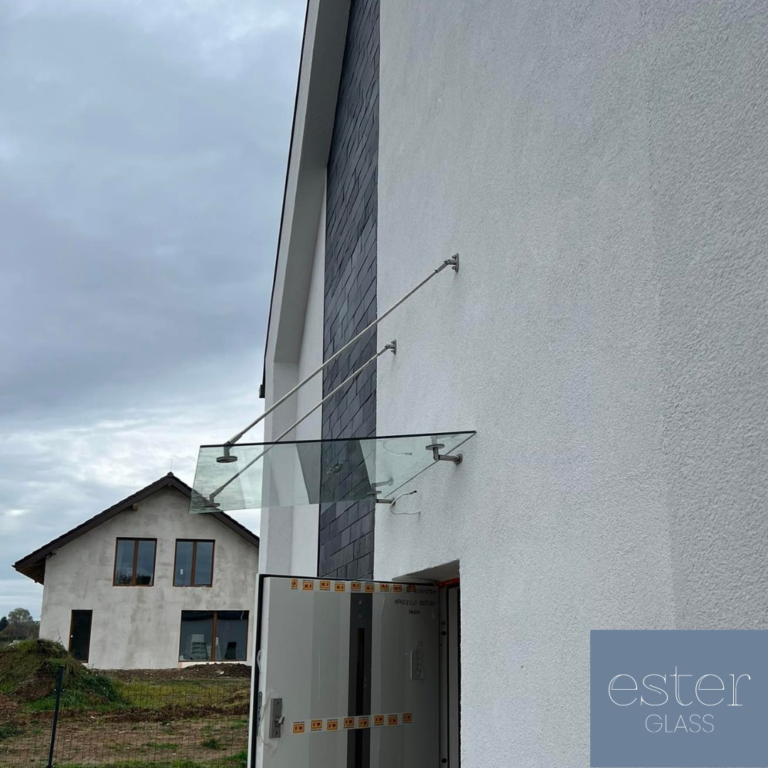 Szklany daszek nad wejściem do domu z białym tynkiem i elementami z szarego łupka, wsparty na metalowych prętach, widoczny dom w tle w trakcie budowy, logo firmy w prawym dolnym rogu.